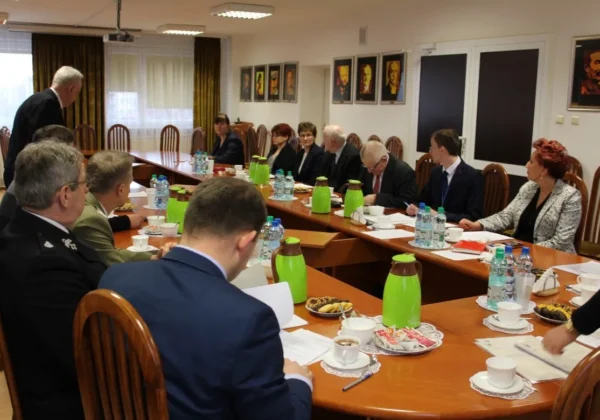 Grupa osób w formalnych ubraniach siedzi przy długim stole w sali konferencyjnej podczas spotkania Komisji Bezpieczeństwa i Porządku, z notatkami, filiżankami kawy, butelkami wody i zielonymi dzbankami na stole.