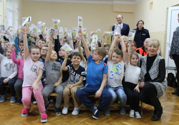 Grupa dzieci wraz z dorosłymi siedzi w szkolnej sali i trzyma w górze opakowania z odblaskami w ramach kampanii „Jesteś widoczny, jesteś bezpieczny”.