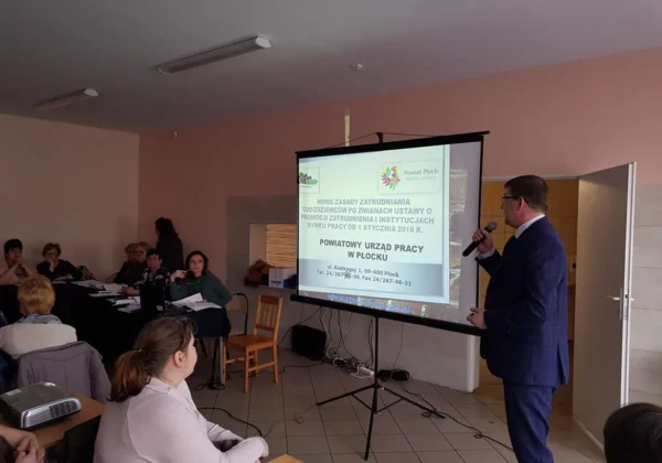 Mężczyzna w garniturze prowadzi prezentację na temat zatrudniania cudzoziemców podczas spotkania informacyjnego w Wyszogrodzie; na ekranie projektora widoczna jest prezentacja Powiatowego Urzędu Pracy w Płocku.