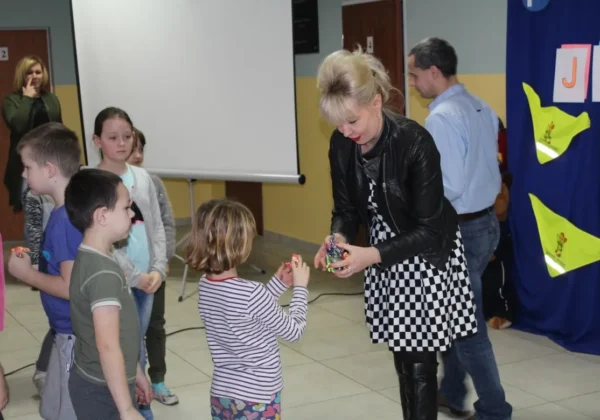 Dzieci stoją w kolejce, dorosła kobieta wręcza im drobny upominek podczas szkolnej kampanii „Jesteś widoczny, jesteś bezpieczny” w SP w Zągotach.