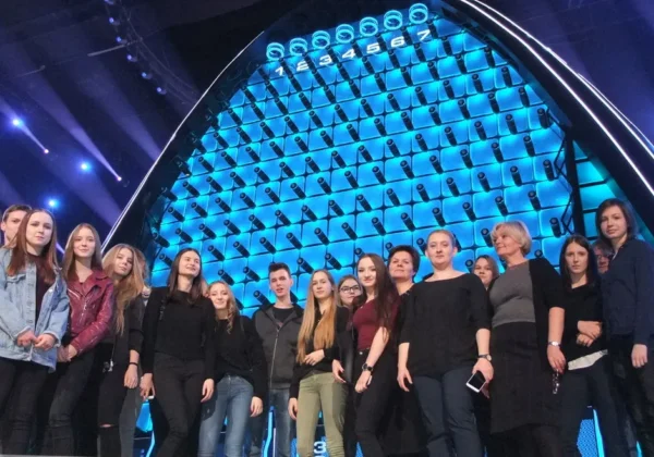 Grupa uczestników stoi na scenie przed imponującą niebieską ścianą The Wall z licznymi panelami i efektownym oświetleniem.