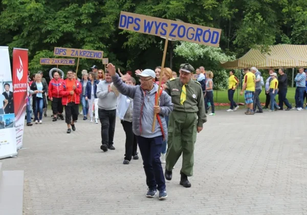 Grupa osób maszeruje po wybrukowanym placu z banerem „DPS WYSZOGRÓD” na czele, podczas akcji pomocy dla chorych, starszych i dzieci.