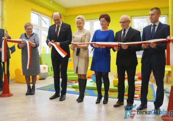 Grupa samorządowców w eleganckich strojach przecina czerwoną i białą wstęgę podczas uroczystego otwarcia Żłobka Samorządowego w Nowym Grabiu, w kolorowym wnętrzu wypełnionym zabawkami.