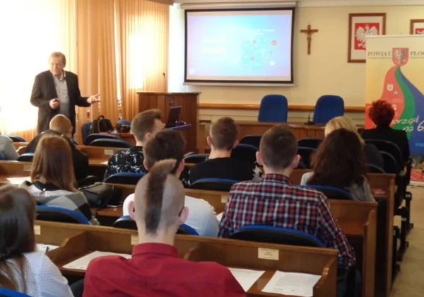 Mężczyzna prowadzi prezentację w sali konferencyjnej przed grupą słuchaczy; za nim projektor wyświetlający slajd i baner Powiatu Płockiego.
