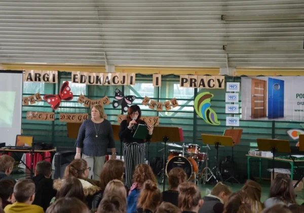 Dwie kobiety prowadzą prezentację na scenie podczas XII Targów Edukacji i Pracy w Małej Wsi; przed sceną siedzi grupa uczniów, a w tle wiszą dekoracje z motylami i baner edukacyjny.