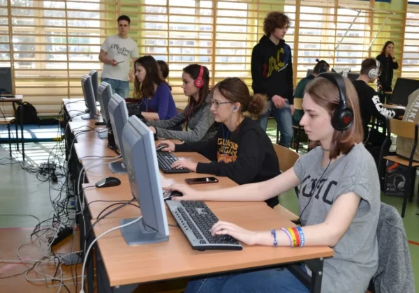 Grupa młodych graczy siedzi przy stanowiskach komputerowych z monitorami i słuchawkami, grając w turnieju League of Legends w szkolnej hali.
