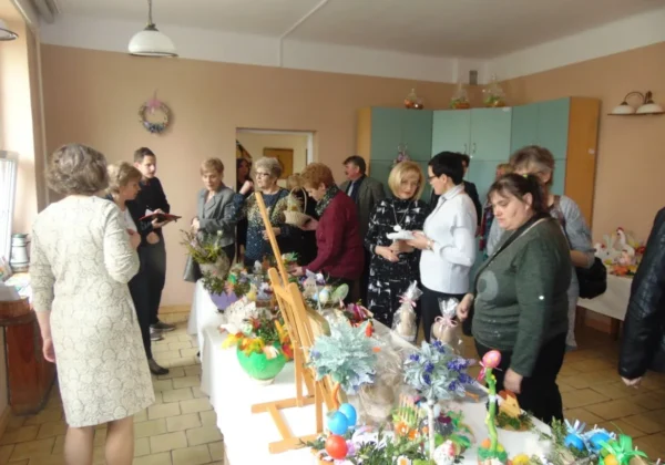 Grupa mieszkańców Zakrzewa ogląda dekoracje i rękodzieła rozłożone na stołach podczas świątecznego spotkania.