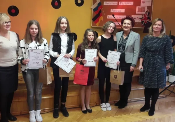 Grupa dziewcząt i dorosłych na scenie podczas Powiatowego Festiwalu Piosenki Obcojęzycznej, trzymających dyplomy i upominkowe torby.