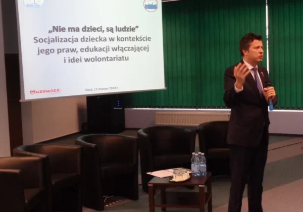 Mężczyzna w garniturze przemawia podczas konferencji; na ekranie widnieje slajd z hasłem „Nie ma dzieci, są ludzie” dotyczącym socjalizacji, edukacji włączającej i idei wolontariatu.