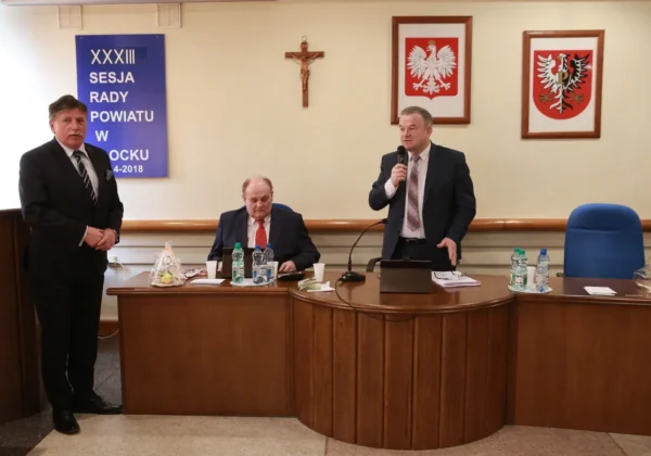 Trzech mężczyzn w garniturach podczas XXXIII sesji Rady Powiatu; jeden stoi po lewej, drugi zabiera głos z mikrofonem po prawej, a trzeci siedzi przy okrągłym stole, w tle krzyż, herby i baner informujący o sesji.