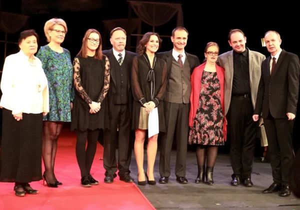 Grupa dziewięciu osób w formalnych strojach stoi na scenie Teatru Płockiego na czerwonym dywanie podczas prapremiery.
