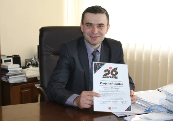 Mężczyzna w garniturze siedzi przy biurku w biurze i prezentuje dokument z napisem „26 Finał”.