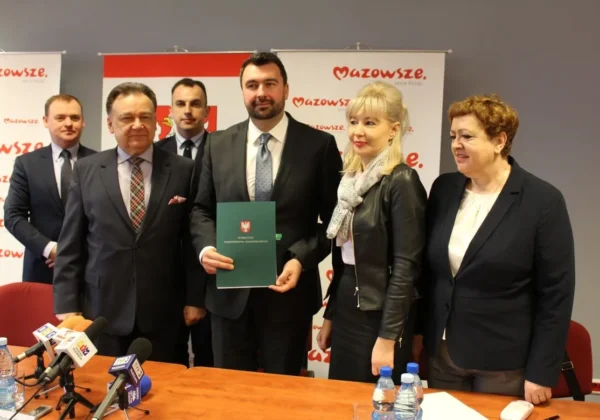 Grupa pięciu osób w garniturach stoi na konferencji prasowej, a mężczyzna w środku trzyma zieloną teczkę z godłem, na tle widoczne banery Mazowsza.