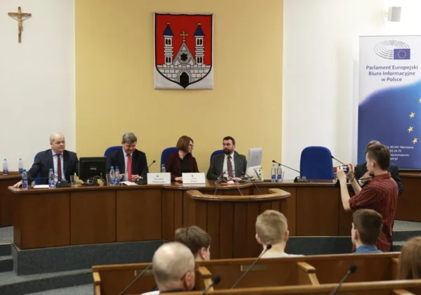 Panel dyskusyjny z posłami do Parlamentu Europejskiego w sali konferencyjnej, siedzący za długim stołem z mikrofonami i wodą, w tle widoczny herb i baner Parlamentu Europejskiego.