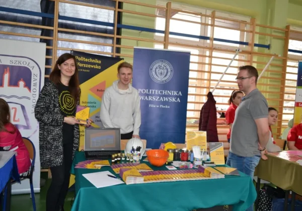 Stoisko Politechniki Warszawskiej (filia w Płocku) podczas III Targów Edukacyjno‑Zawodowych „Bieg do przyszłości” z rozmówcami, laptopem, materiałami promocyjnymi i gadżetami na stole oraz bannerami uczelni w tle.