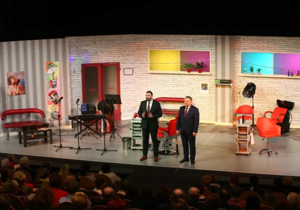 Na scenie Powiatu Płockiego dwóch mężczyzn w garniturach przemawia do widowni podczas spektaklu „Szalone nożyczki” z okazji Dnia Kobiet; w tle widać czerwone fotele, instrumenty i kolorową scenografię.