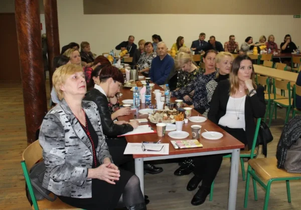 Grupa uczestników konferencji edukacyjnej w Radzanowie siedzi przy długim stole, mając przed sobą notatniki, napoje i przekąski.
