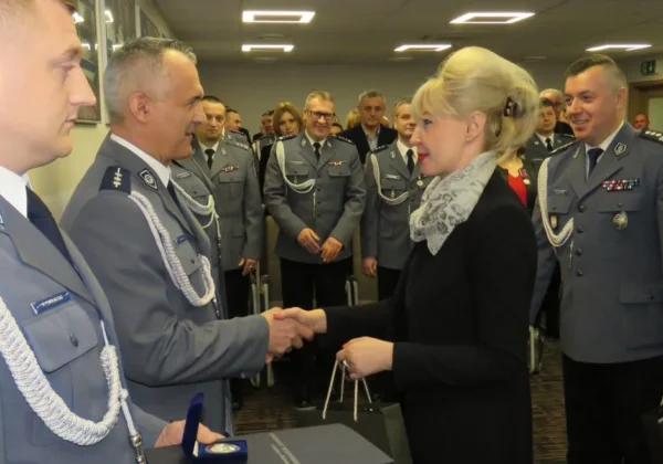 Osoba w czarnym stroju wręcza odznaczenie policjantowi w mundurze, na tle innych funkcjonariuszy podczas uroczystości konkursu Najpopularniejszego Dzielnicowego Roku.