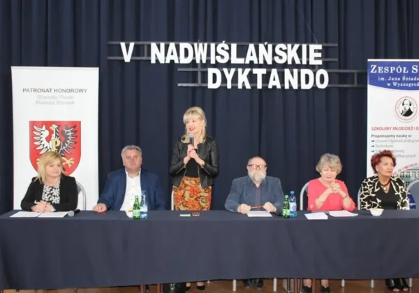 Panel dyskusyjny podczas V Nadwiślańskiego Dyktanda — pięć osób siedzi za stołem na scenie, a jedna z nich stoi i przemawia, w tle widoczny jest duży napis "V Nadwiślańskie Dyktando".