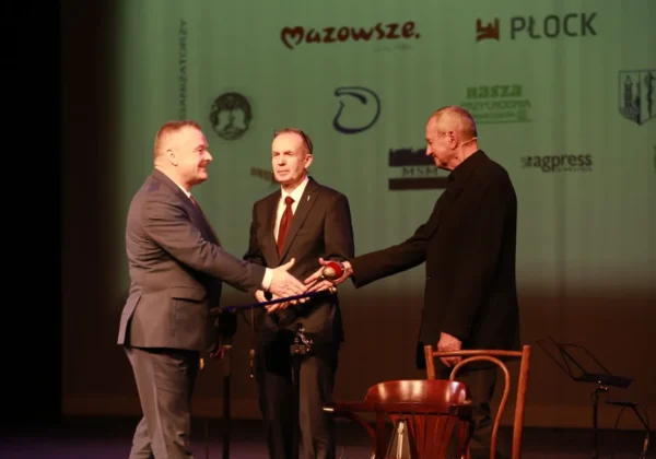 Trzech mężczyzn na scenie podczas jubileuszu 50-lecia Płockiego Towarzystwa Przyjaciół Teatru, dwóch ściska sobie dłonie, trzeci obserwuje, a w tle widoczny banner z logotypami.