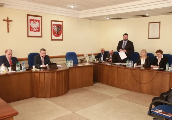 Grupa urzędników siedzi przy długim, zakrzywionym stole podczas formalnego posiedzenia Rady Powiatu Płockiego; stojący mężczyzna prezentuje dokument.