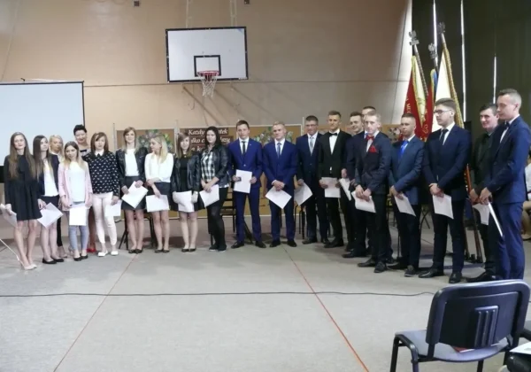 Grupa maturzystów w eleganckich garniturach i sukienkach stoi w rzędzie na sali podczas pożegnania w Gąbinie, trzymając dyplomy.