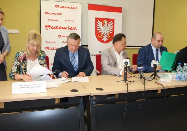Grupa urzędników samorządowych podpisuje dokumenty podczas konferencji prasowej, na tle banerów Mazowsze i polskiego godła.