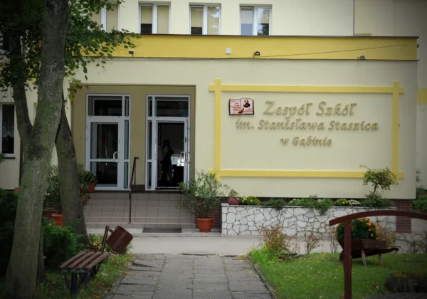 Wejście do Zespołu Szkół im. Stanisława Staszica w Gąbinie, żółto-beżowy budynek z widocznym napisem na ścianie, drzwi wejściowe i otoczenie z kwiatami oraz ławkami.