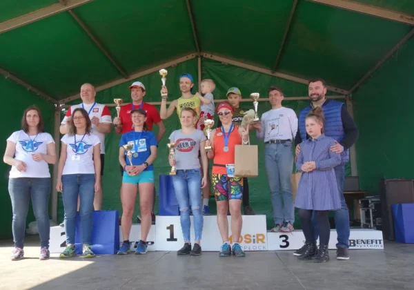 Grupa biegaczy i organizatorów stoi na podium z trofeami i medalami podczas biegu „Razem dla autyzmu” w malowniczych plenerach powiatu płockiego.