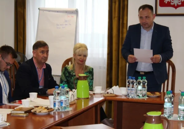 Grupa urzędników przy stole na posiedzeniu Powiatowego Zespołu Zarządzania Kryzysowego omawiająca pożary w Gminie Nowy Duninów, podczas gdy jeden mężczyzna stoi i odczytuje dokumenty.