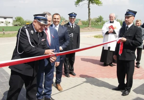 Uroczystość Dnia Strażaka w Wrogocinie: strażacy w galowych mundurach przecinają czerwoną wstęgę podczas ceremonii, w towarzystwie duchownego i innych gości.