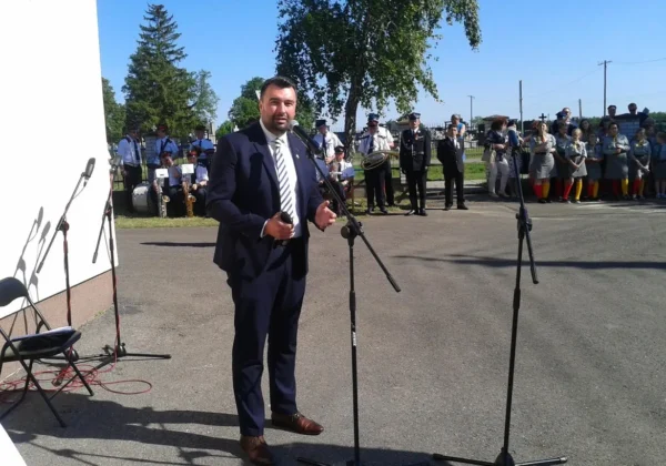 Mężczyzna w garniturze wygłasza przemówienie przy mikrofonie podczas obchodów Dnia Strażaka w gminie Słupno, na tle orkiestry i strażaków w mundurach.