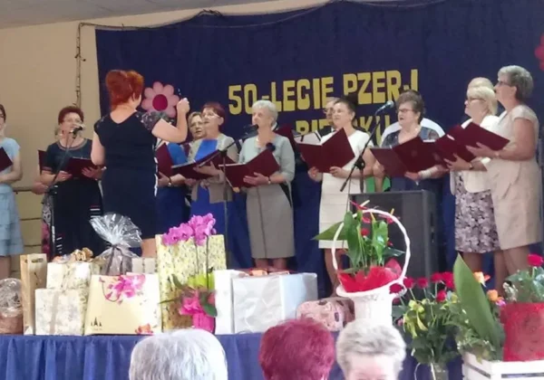 Chór seniorek z Gminy Bielsk na scenie podczas złotego jubileuszu, prowadzony przez dyrygentkę, z prezentami i kwiatami ustawionymi na pierwszym planie.
