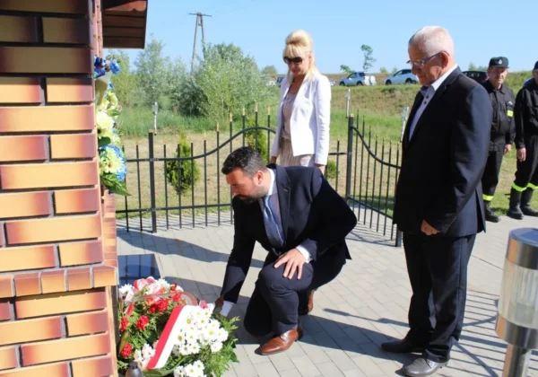 Mężczyzna w garniturze klęka i składa kwiaty przy pomniku upamiętniającym powódź; obok stoją kobieta w białej marynarce i inni oficjele, a w tle widoczni są strażacy.