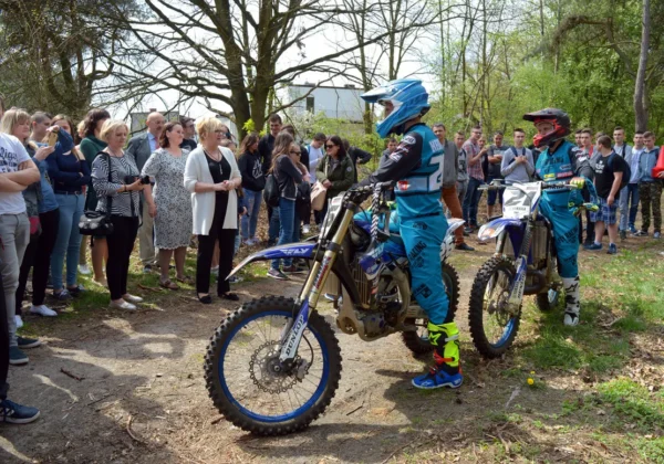 Dwa motocyklistyczne zawodniki w niebieskich kombinezonach i kaskach stoją na szutrowej drodze, obserwowani przez tłum widzów podczas wydarzenia Motograwitacja – Bezpieczna Droga 2018.