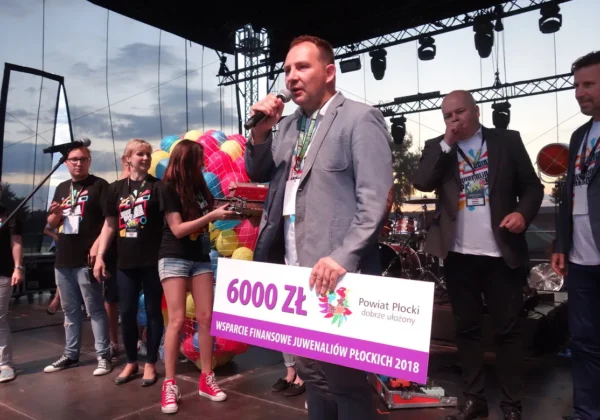 Mężczyzna w garniturze trzyma mikrofon i prezentuje duży czek na kwotę 6000 zł podczas Juwenaliów Płockich 2018, na scenie w otoczeniu innych uczestników i balonowej dekoracji.