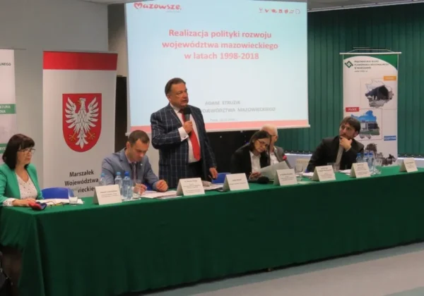 Panel dyskusyjny w sali konferencyjnej z kilkoma osobami siedzącymi przy zielonym stole, z prowadzącym przemawiającym do mikrofonu na tle prezentacji o rozwoju Mazowsza.
