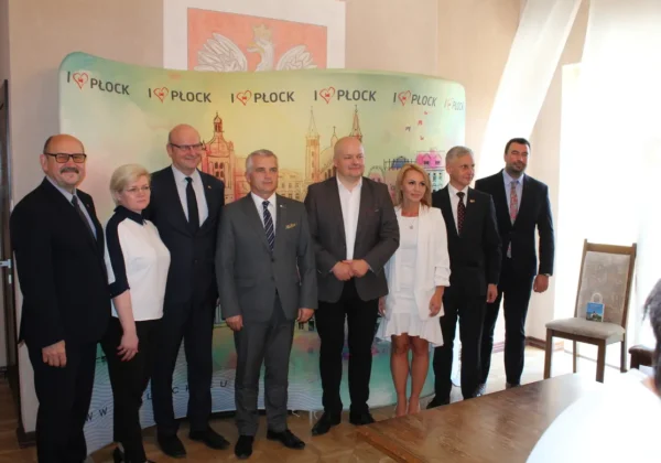 Grupa ośmioosobowa delegacji parlamentu litewskiego w formalnych strojach stoi przed kolorowym banerem „I ♥ Płock” podczas roboczej wizyty w ziemi płockiej.
