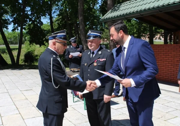 Na zewnątrz budynku strażak w mundurze i urzędnik w garniturze ściskają dłonie, przekazując dokumenty podczas ceremonii nominacyjnej Starostwa Powiatowego w Płocku do Strażackich Oscarów.