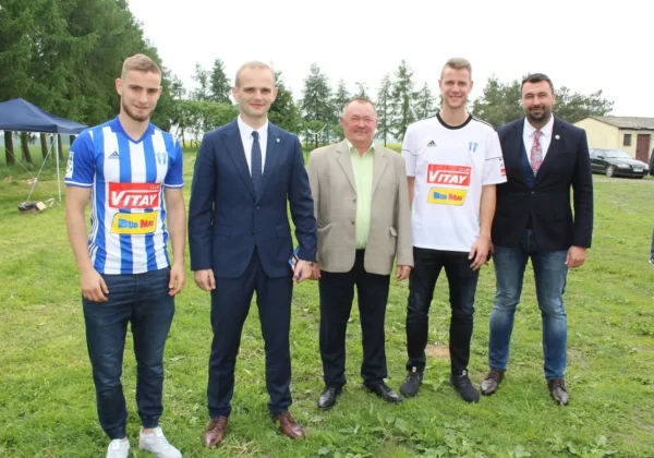 Pięciu mężczyzn stoi na trawniku podczas Pikniku Rodzinnego w Warsztacie Terapii Zajęciowej w Męczeninie, dwóch w koszulkach piłkarskich, pozostali w marynarkach, a w tle widać namioty i drzewa.