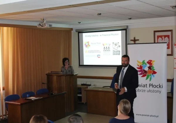 Mężczyzna w garniturze wygłasza przemówienie podczas prezentacji w sali konferencyjnej; obok niego stoi kobieta przy podium, na ekranie widać logotypy, a z boku stoi baner Powiatu Płockiego.