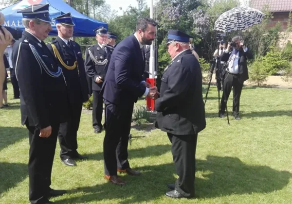 Uroczystość jubileuszowa strażaków Ochotników w Powiecie Płockim: mężczyzna w garniturze wręcza odznaczenie starszemu strażakowi w mundurze, podczas gdy pozostali strażacy i fotografowie stoją w tle.