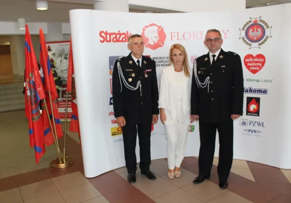 Trójka osób stoi na tle ścianki z logotypami i flagami; dwóch strażaków w czarnych mundurach z odznaczeniami i srebrnymi sznureczkami na ramionach stoi obok kobiety w białym garniturze.