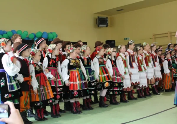 Dzieci w tradycyjnych, kolorowych strojach ludowych stoją w rzędzie na sali gimnastycznej podczas jubileuszu 25-lecia Szkoły Podstawowej Specjalnej nr 24 w Płocku.