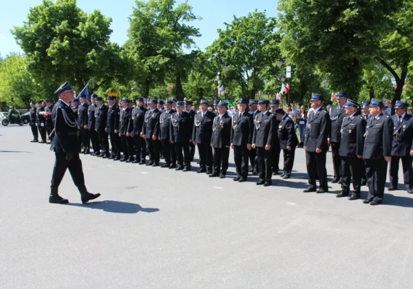 Grupa strażaków Ochotniczej Straży Pożarnej w Gąbinie stoi w rzędzie na ulicy podczas obchodów Dnia Strażaka i przekazania nowego samochodu ratowniczo-gaśniczego, a dowódca przechodzi na pierwszy plan.