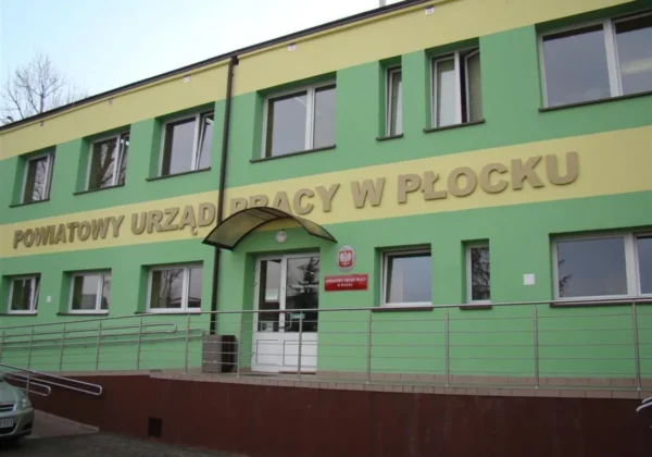 Dwupiętrowy budynek z zielono‑żółtą elewacją oznaczony napisem Powiatowy Urząd Pracy w Płocku, z wejściem pod daszkiem i rampą dla niepełnosprawnych.