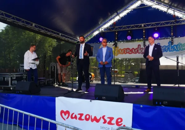 Trzech mężczyzn w garniturach stoi na scenie podczas XIV Jarmarku Norbertańskiego w Bodzanowie, z mikrofonami i sprzętem muzycznym w tle.