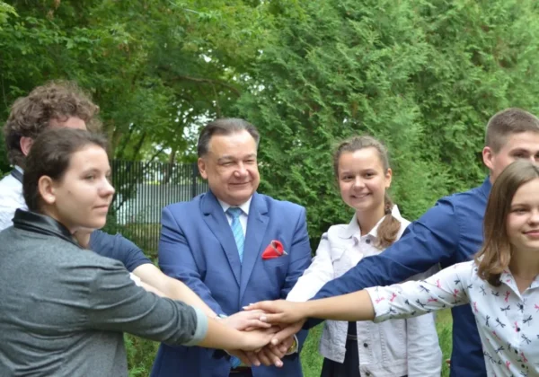 Grupa młodych ludzi i urzędnik w niebieskim garniturze łączą dłonie w geście jedności podczas inauguracji stypendiów Marszałka dla najzdolniejszej młodzieży z Mazowsza, na tle zieleni.