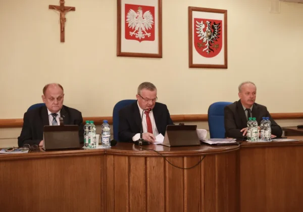 Trzech mężczyzn w garniturach siedzi przy drewnianym stole w sali obrad podczas XXXVI sesji Rady Powiatu; centralny mężczyzna mówi do mikrofonu, a na ścianie wiszą krzyż i dwa herby.