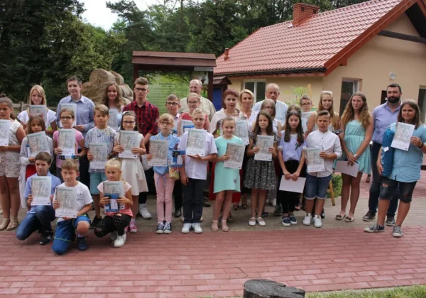 Grupa uczniów i opiekunów stoi na zewnątrz przed budynkiem, trzymając certyfikaty, podczas podsumowania konkursu ekologicznego dla dzieci i młodzieży z powiatu płockiego – II etap.